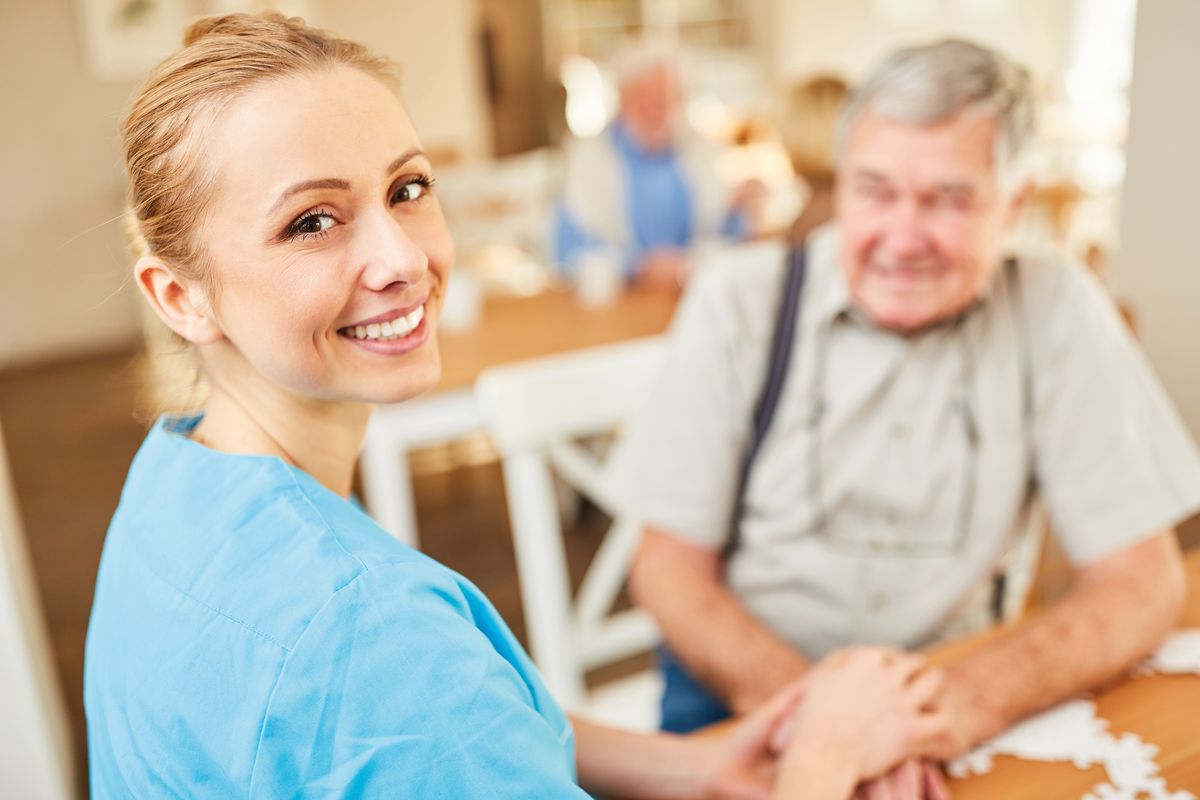 Eine Pflegerin schaut lächelnd in die Kamera und hinter ihr sitzt leicht unscharf ein alter Mann der auch sehr zufrieden wirkt.