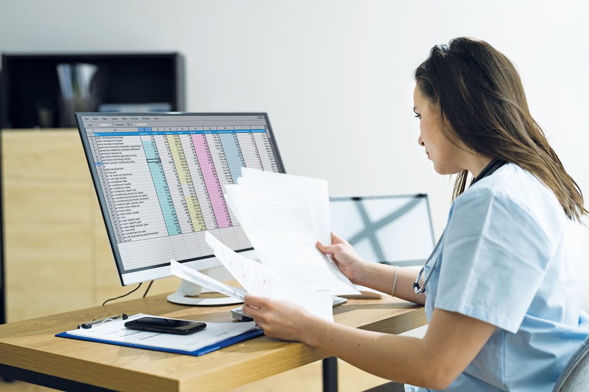 Eine Person aus dem Gesundheitswesen arbeitet an einem Computer und hat dabei verschiedene Bögen Papier in der Hand.