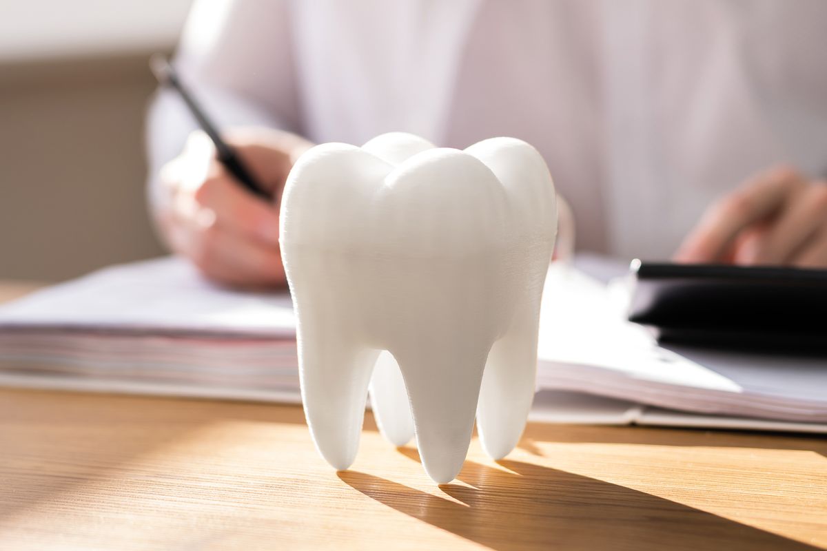 Ein großer Zahn steht auf einem Tisch vor einem Zahnarzt, der gerade etwas aufschreibt.